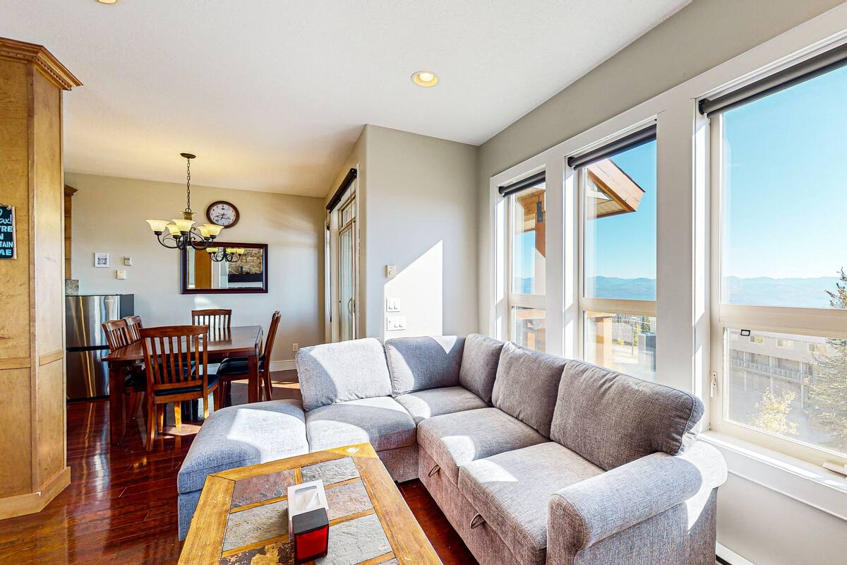 A modern living room with a sectional sofa, a dining area with wooden chairs, and large windows offering a view.