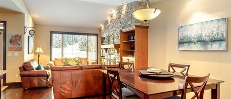 Un comedor con mesa y sillas de madera, chimenea de piedra y vistas al exterior a través de amplios ventanales.
