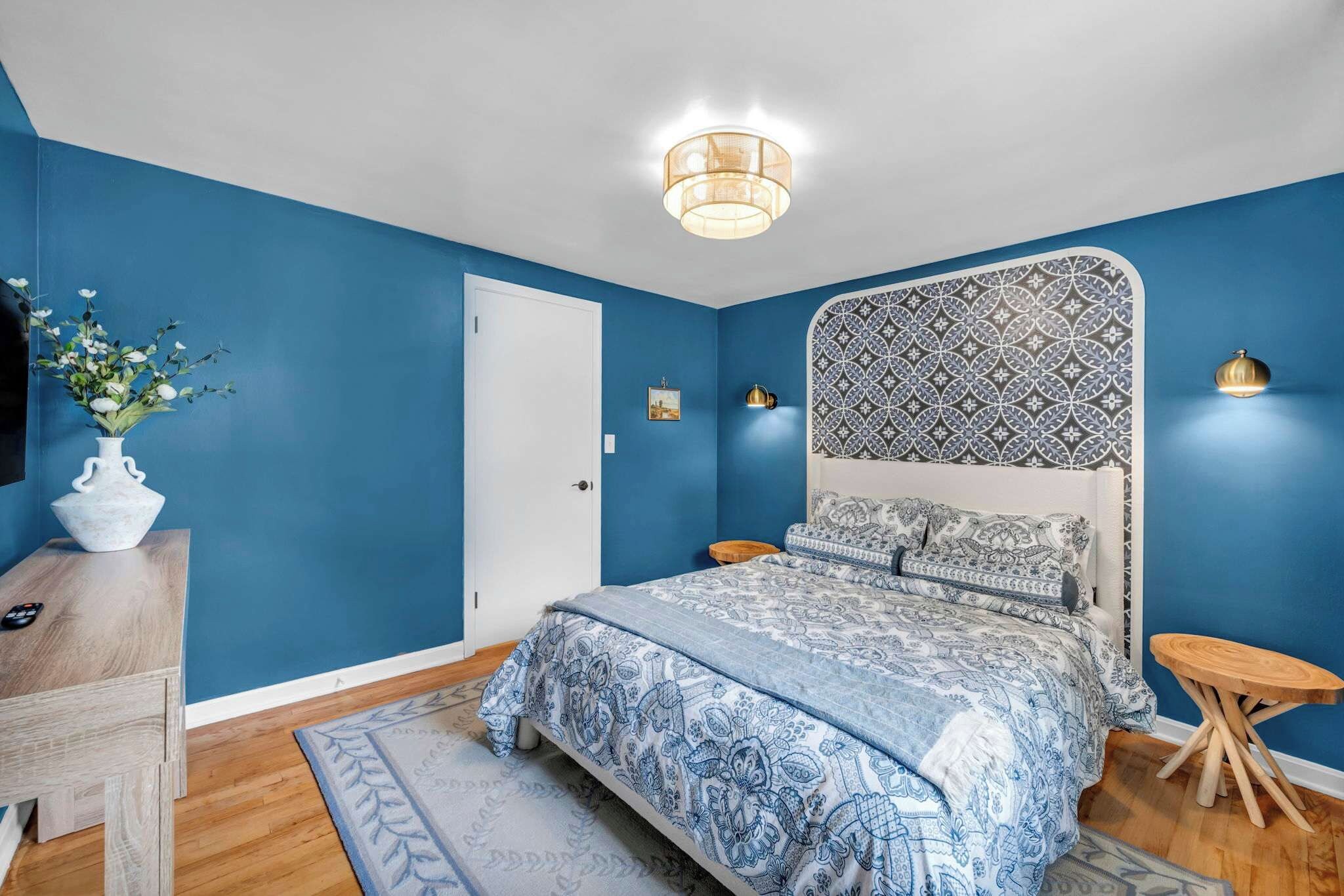 Quarto com cama, cômoda, mesa de cabeceira, abajur e um vaso com flores.