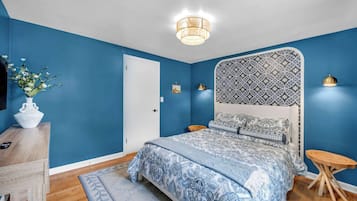 Quarto com cama, cômoda, mesa de cabeceira, abajur e um vaso com flores.