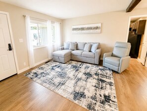 Ein Wohnzimmer mit einem Ecksofa, einem hellblauen Sessel, einem gemusterten Teppich und einer durchgehende offene Tür sichtbaren Küche.