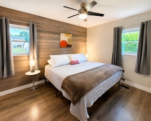 Un dormitorio con cama, ventilador de techo, mesita de noche, lámpara y ventana con cortinas.