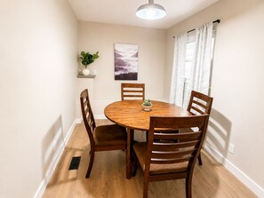 Een eethoek met een ronde houten tafel, vier houten stoelen, een potplant, een wandplank en een schilderij.