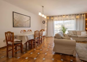 Un comedor con una mesa y sillas, un sofá, un cuadro grande y un ventanal con cortinas.