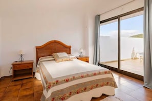 Un dormitorio con cama de madera, dos mesitas de noche con lámparas, un ventanal con vistas al paisaje y una silla de balcón.