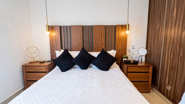 A neatly made bed with a white quilt, two black pillows, and a wooden headboard with vertical slats. There are two bedside tables with lamps, a small round mirror, and a decorative metal piece on the left table.