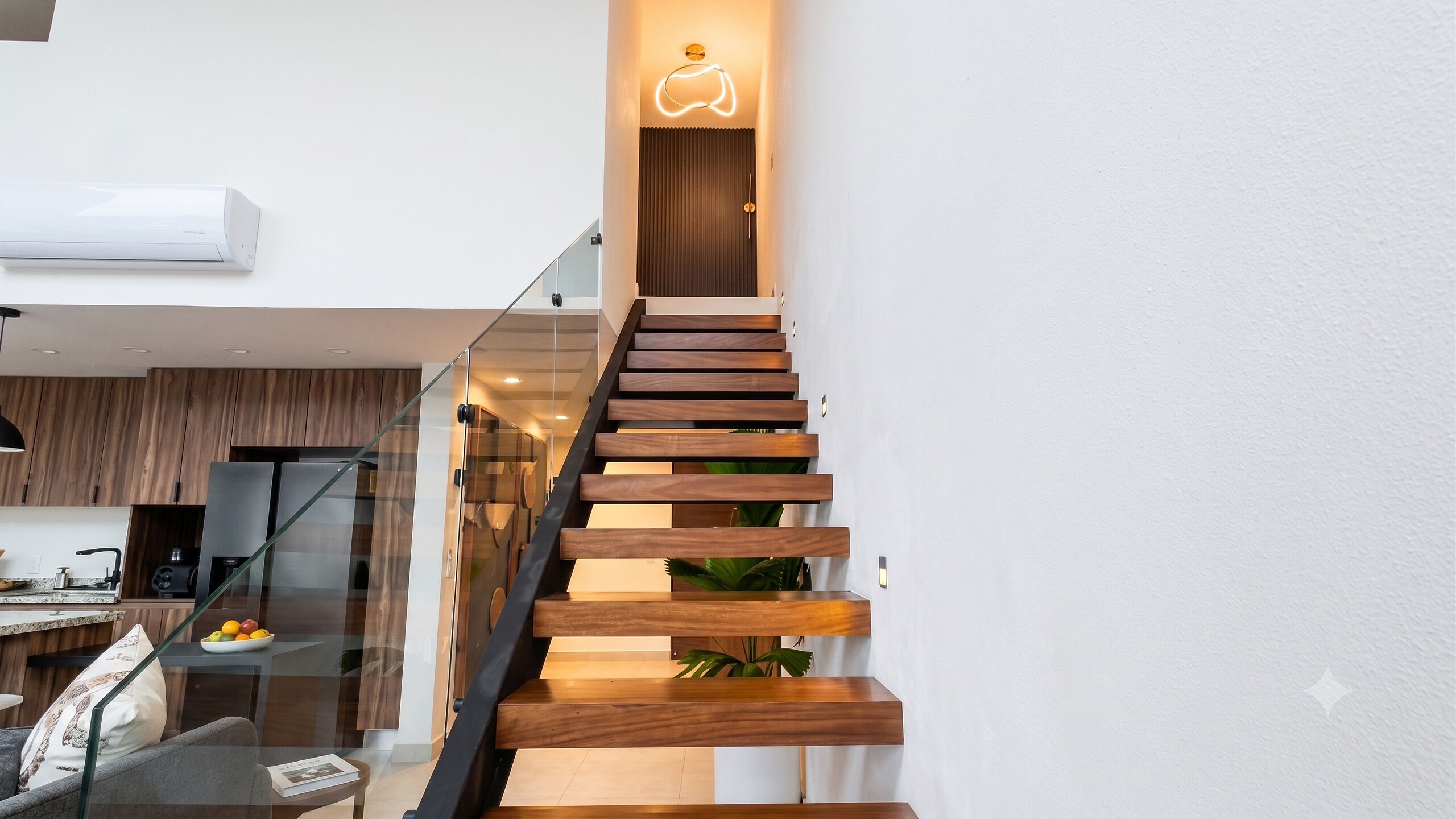 Una escalera moderna con escalones de madera y barandillas de vidrio, que conduce a una habitación con cocina y comedor.