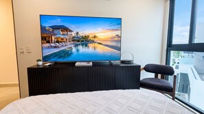 A bedroom with a large TV displaying a resort scene, a bed, a chair, and a window with a view of buildings.