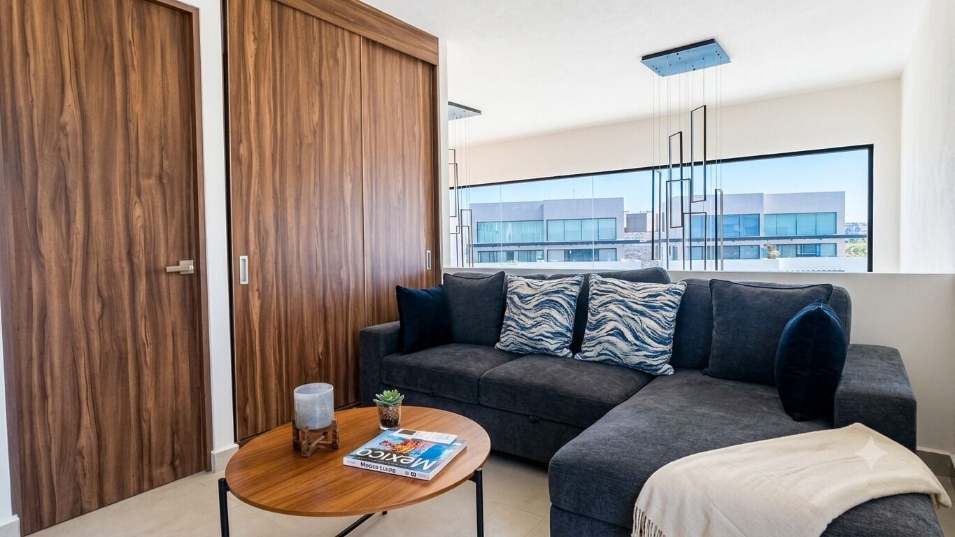 Un salon moderne avec un canapé gris, une table basse en bois et un magazine. Une grande fenêtre offre une vue sur les bâtiments à l’extérieur.