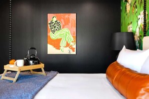 Ein Zimmer mit einem Bett, einem Holftablett mit Wasserkocher, Tasse und einem Muffin, einem Bild an der Wand und einer Lampe auf einem Nachttisch.