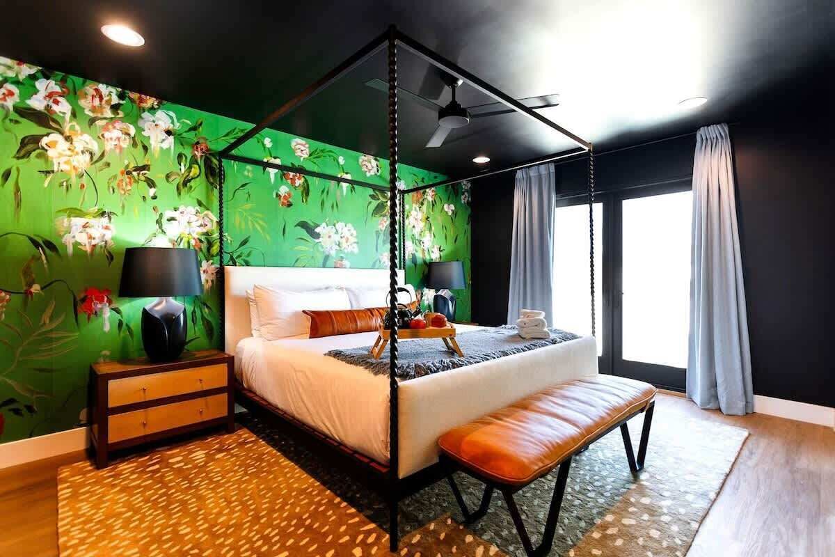 A four-poster bed with a canopy, a bedside table with lamps, a bench, a tray with fruits, and a large window with curtains.