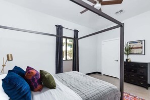 Ein Schlafzimmer mit Himmelbett, Deckenventilator, einem Fenster mit Vorhängen, einer schwarzen Kommode und einem gerahmten Bild an der Wand.