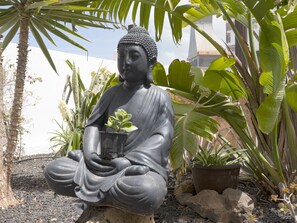 Eine sitzende Buddha-Statue mit einem Blumentopf, umgeben von tropischen Pflanzen und Steinen.