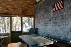 A cabin interior with a wooden ceiling, a table, benches, and a view of a snowy forest through large windows.