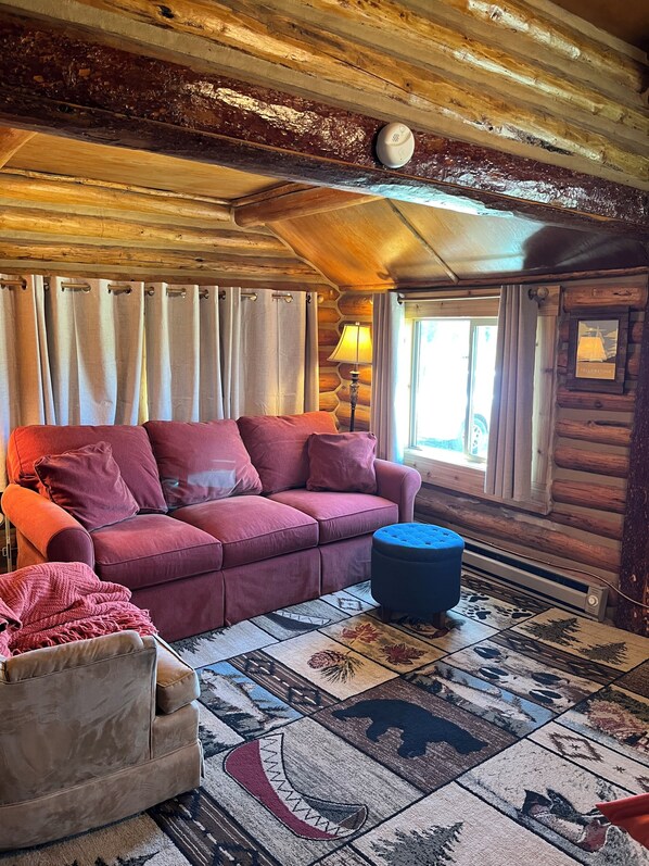 Ein rustikaler Innenraum einer Blockhütte mit einem roten Sofa, einem blauen Hocker und einem gemusterten Teppich.