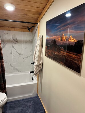 Ein Badezimmer mit einer Badewanne, einer Toilette, Marmorfliesen, einer blauen Fußmatte, einem Handtuch auf einem Haken und einem großen Landschaftsbild an der Wand.