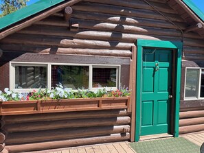 Eine Blockhütte mit grüner Tür, Fenstern mit Vorhängen und einem Blumenkasten mit verschiedenen Pflanzen.
