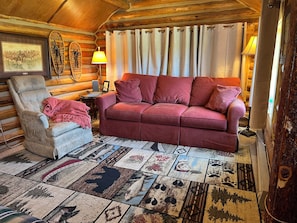 Ein rustikales Blockhaus-Interieur mit einem roten Sofa, einem gepolsterten Sessel, einem gemusterten Teppich und einer Holzbalkendecke.