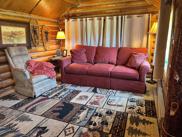 Ein rustikales Blockhaus-Interieur mit einem roten Sofa, einem gepolsterten Sessel, einem gemusterten Teppich und einer Holzbalkendecke.