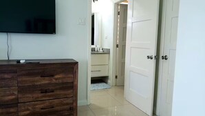 Una habitación con un aparador de madera, un televisor de pantalla plana, una puerta blanca y un baño con espejo y lavamanos.
