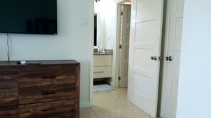 Ein Zimmer mit Holzkommode, Flachbildfernseher, weißer Tür und einem Badezimmer mit Spiegel und Waschbecken.