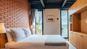 Um quarto com parede de tijolos, uma cama com almofadas, uma mesinha de cabeceira, um guarda-roupa de madeira e uma janela com cortinas.