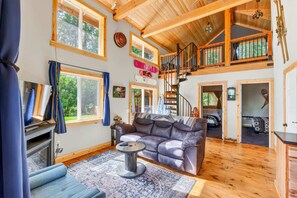 A spacious living room with a high ceiling, wooden beams, a fireplace, a sofa, and a staircase leading to an upper level. - Mt Hood Cabin, Hot Tub, Fire Pit, Dog-Friendly (Sandy)