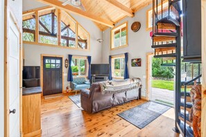 A spacious room with a wooden ceiling, large windows, a sofa, a television, and a staircase. - Mt Hood Cabin, Hot Tub, Fire Pit, Dog-Friendly (Sandy)