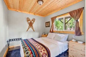 Ein Schlafzimmer mit Holzboden, einem Bett mit bunter Decke, einem Nachttisch, einem Fenster mit Vorhängen und einer wandmontierten Leuchte.