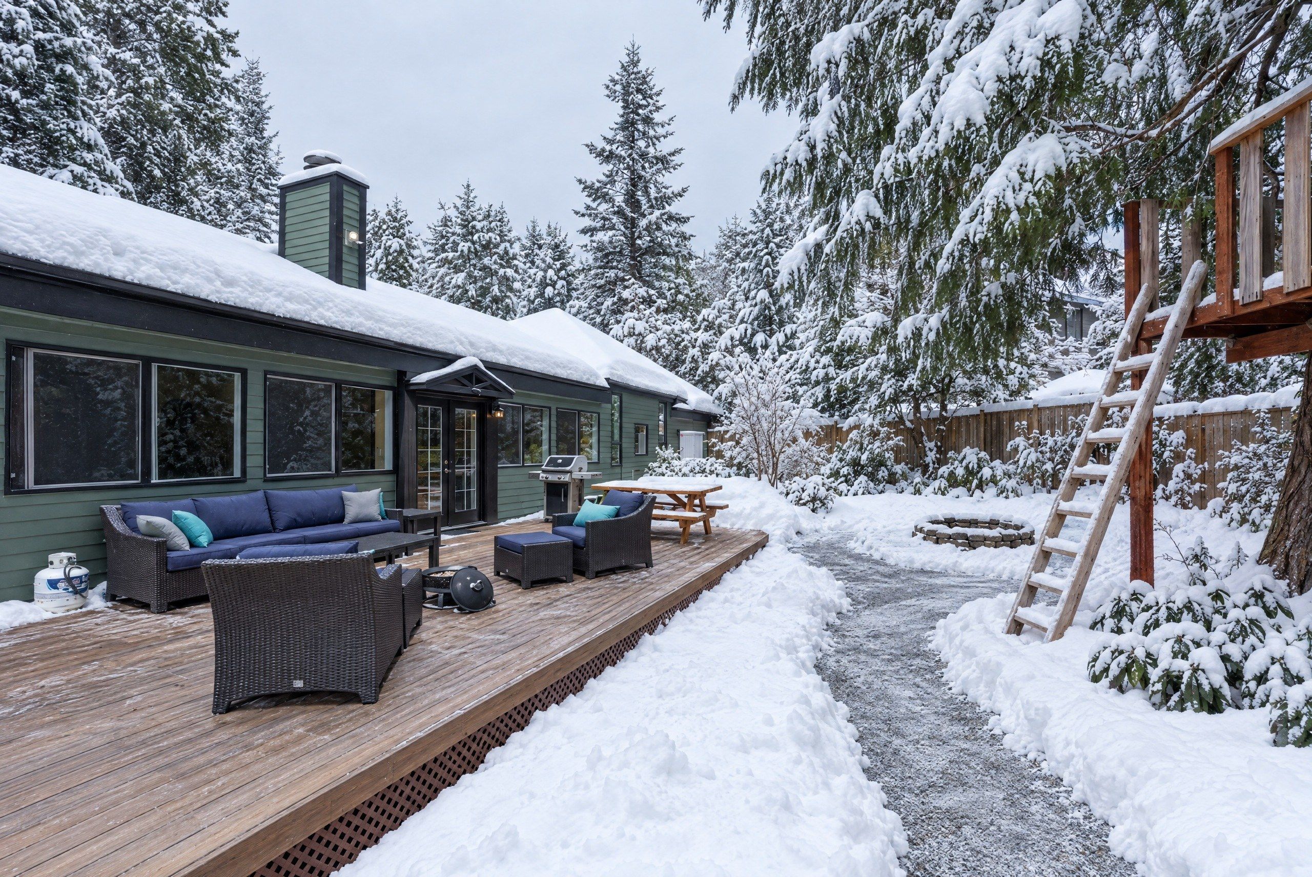 A snow-covered deck with outdoor furniture, a barbecue grill, and a wooden ladder.