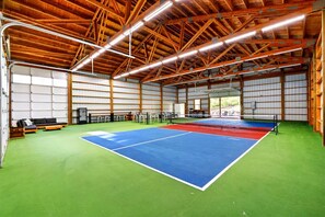 Un centro deportivo interior con cancha de tenis, vigas de madera y piso verde.