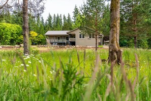 Une maison à deux étages dotée d’une terrasse, entourée d’un paysage naturel avec des arbres et une zone herbeuse.