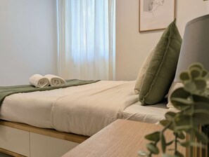 Ein ordentlich bezogenes Bett mit einem grünen Kopfkissen, einem zusammengerollten Handtuch und einem gerahmten Bild an der Wand.