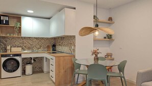 A compact kitchen with a washing machine, a dining area with a glass table and chairs, and a hanging light fixture. - Casa Azahar Apartment Valencia (Valencia)