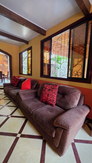 Un salon avec un canapé marron, des coussins aux motifs rouges et noirs, et de grandes fenêtres ornées de barreaux en fer décoratifs.