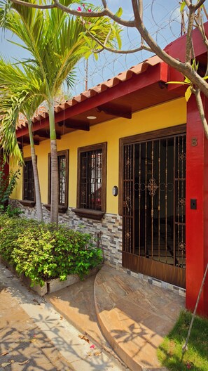 Une maison avec une porte rouge, une fenêtre ornée de barreaux décoratifs, un mur en pierre et un palmier.