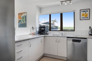 A modern kitchen with white cabinets, a coffee machine, a dishwasher, and a window with a view of palm trees.