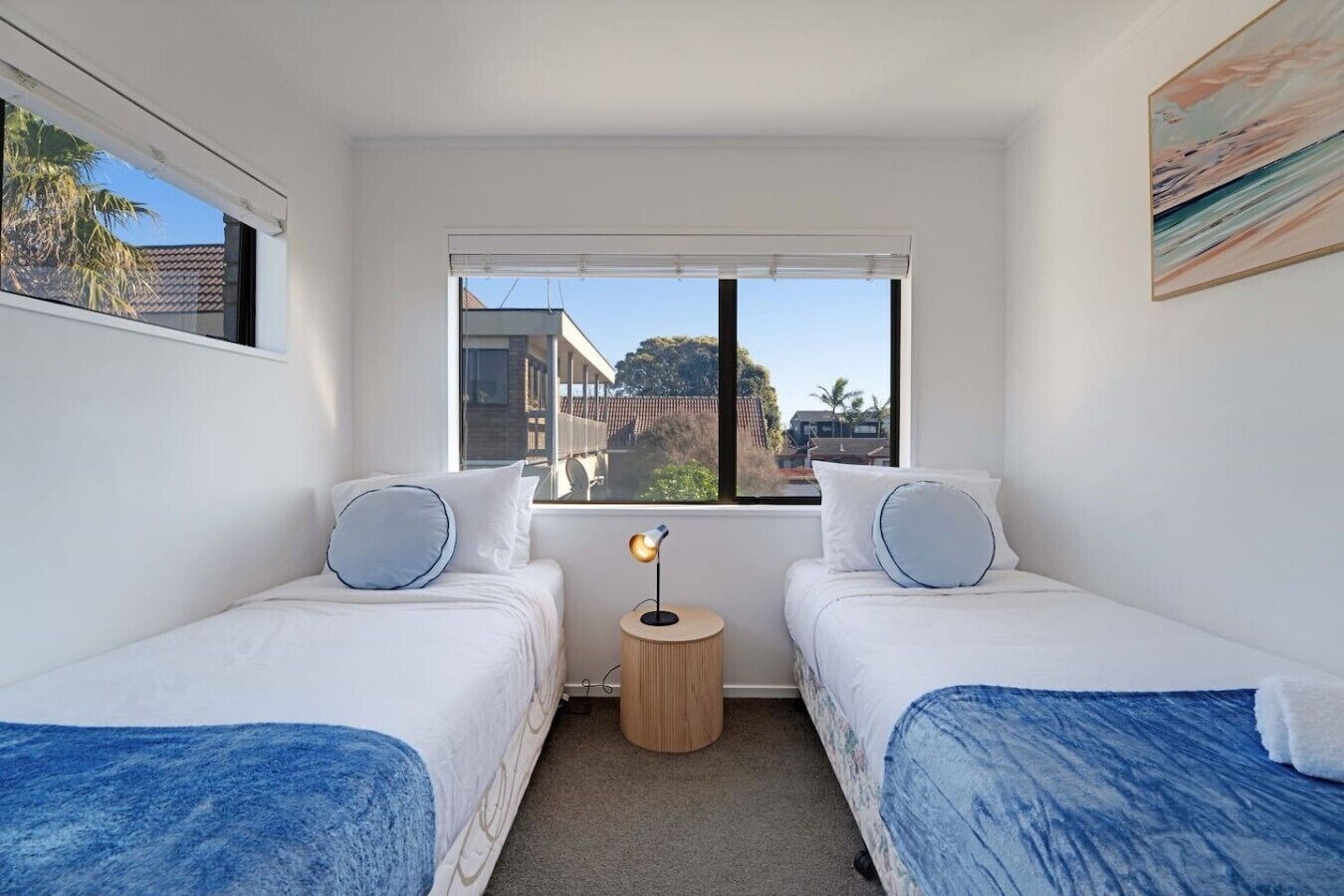 A bedroom with two beds, a small bedside table with a lamp, a window with a view of buildings, and a painting on the wall.