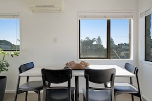 Comedor con una mesa y cuatro sillas, una planta en maceta y vistas a una zona residencial a través de amplios ventanales.