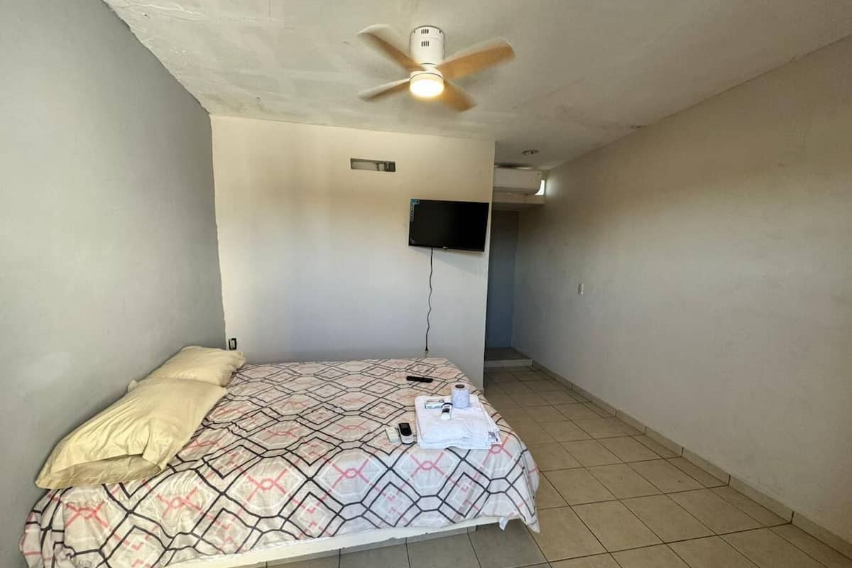 A single bed with a patterned bedspread, a ceiling fan, a mounted television, and a tiled floor.