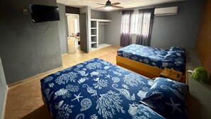 Una habitación con dos camas, ventilador de techo, aire acondicionado, televisor y vista a un pasillo.