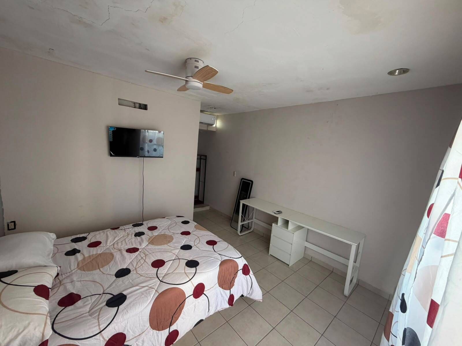 A bedroom with a bed, a desk, a ceiling fan, and a TV mounted on the wall.