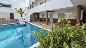 A swimming pool with a palm tree, a building with multiple balconies, and a patio area.