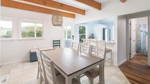 A spacious dining area with a wooden table and chairs, a hanging light fixture, and a view to a kitchen area. - Coastal Getaway with Pool, Spa, Direct Beach Path! (Southern Shores)