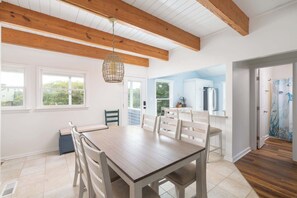 A spacious dining area with a wooden table and chairs, a hanging light fixture, and a view to a kitchen area. - Coastal Getaway with Pool, Spa, Direct Beach Path! (Southern Shores)