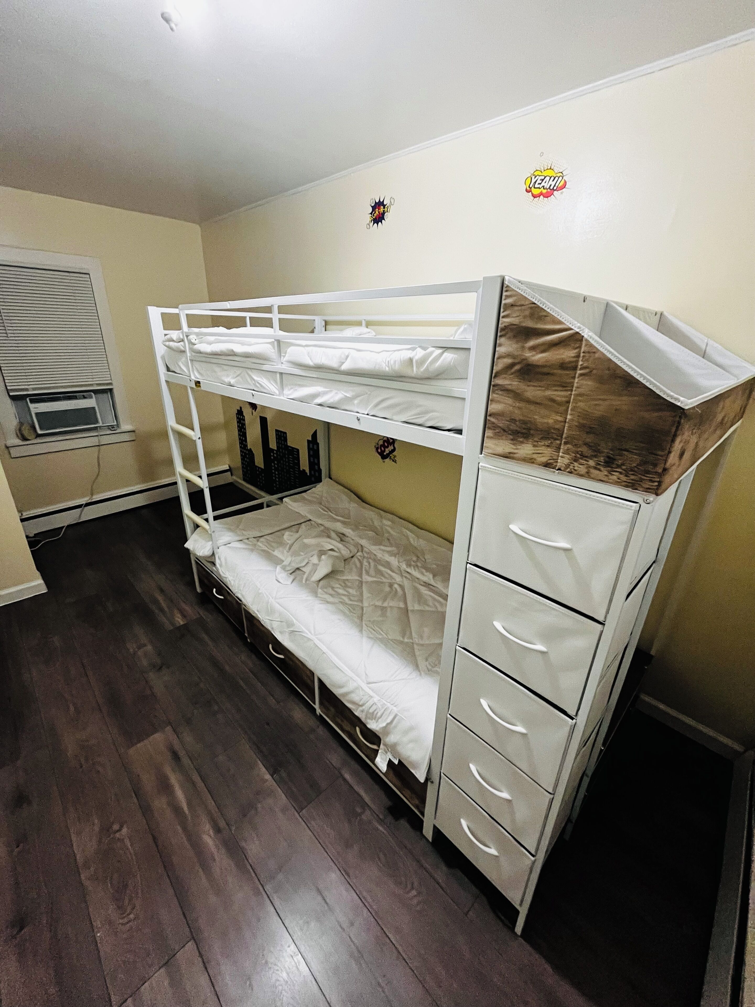 A bunk bed with drawers, a window with blinds, and a wall decoration.