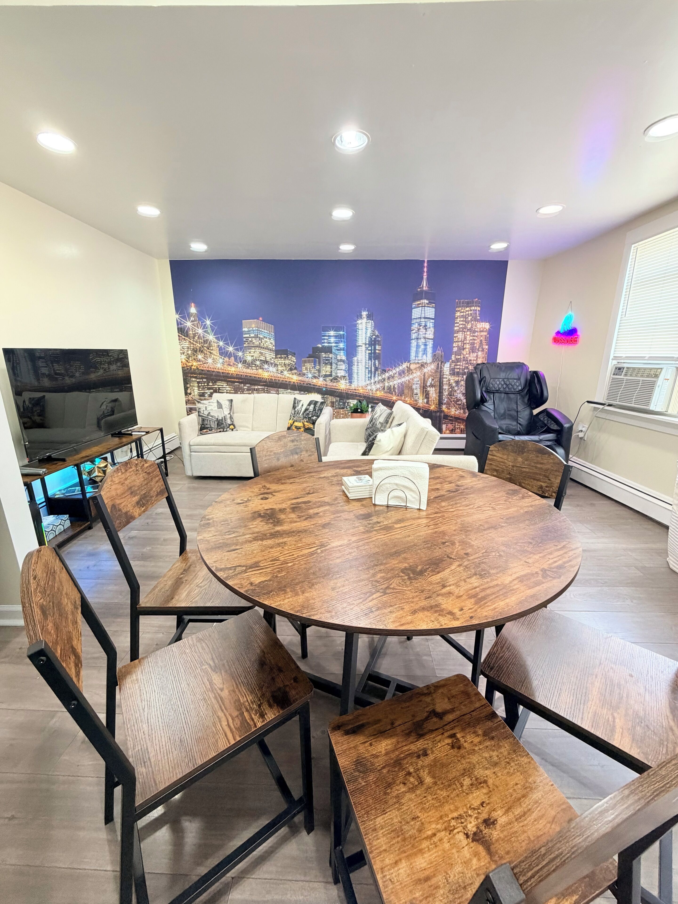 Een moderne woonkamer met een ronde houten tafel, lederen fauteuils, een bank en een muurschildering van een stadsgezicht.