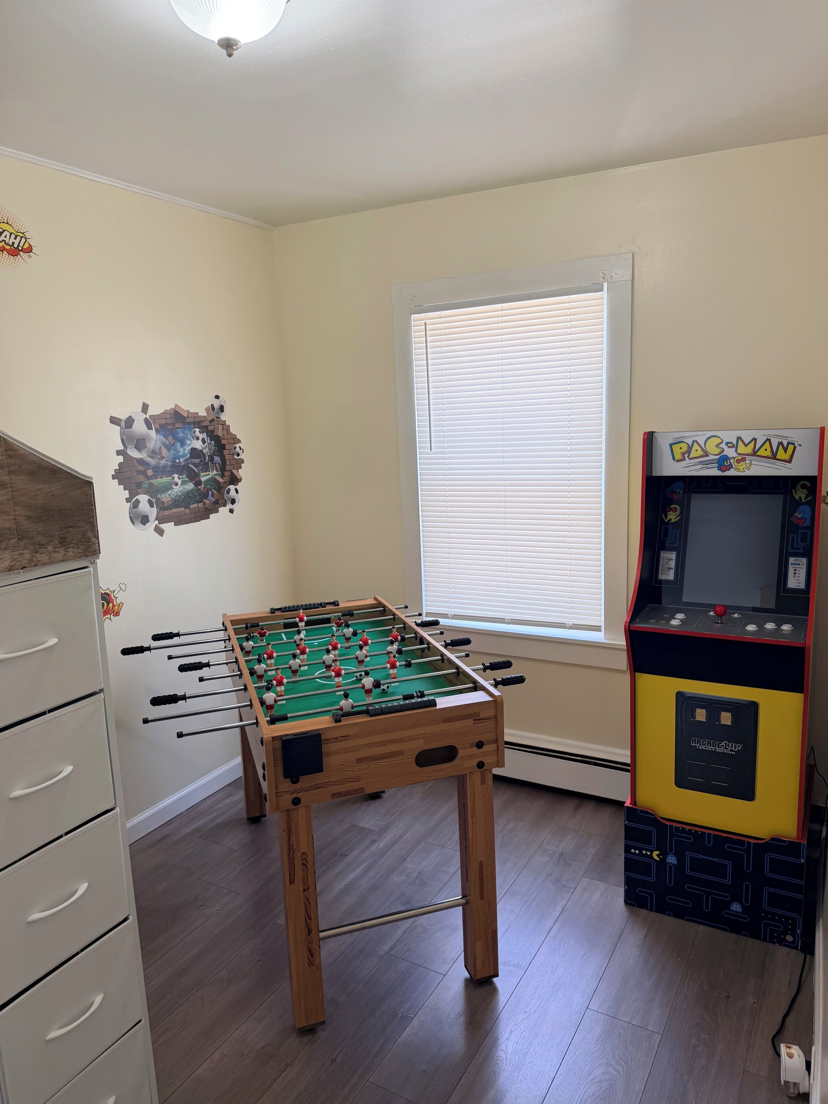Ein Zimmer mit Kicker, Pac-Man Spielautomat und einem Fenster mit Jalousien.