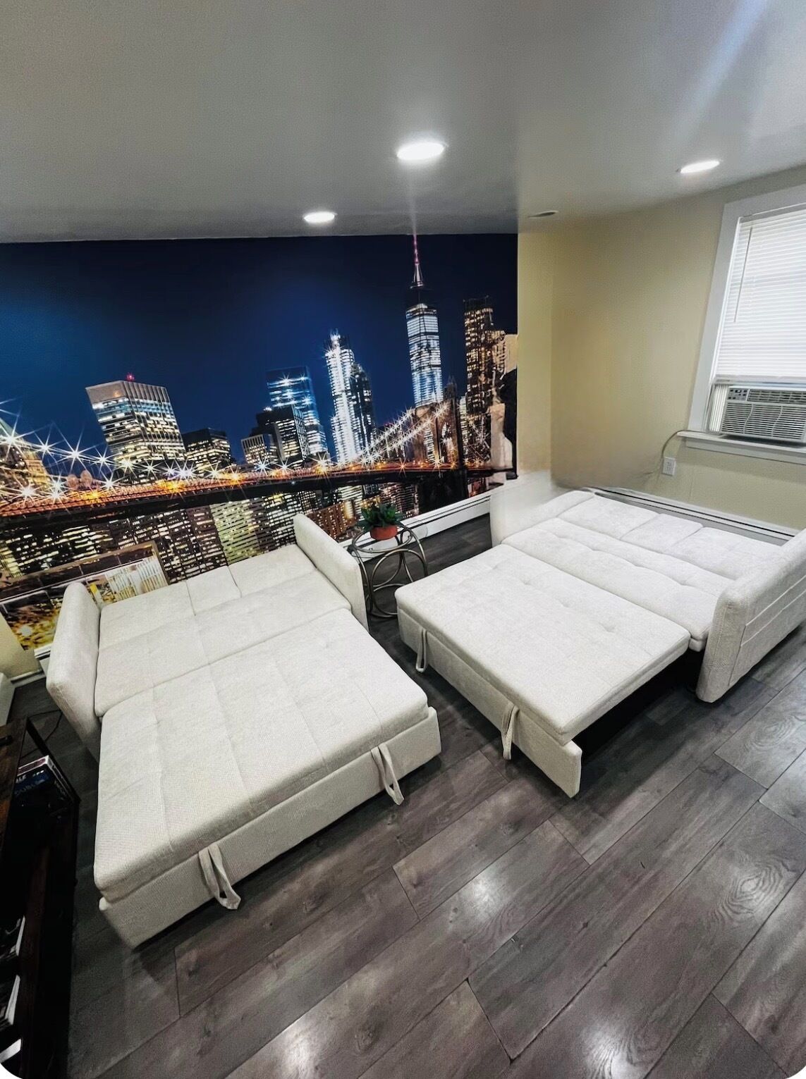 A room with two white beds, a window with blinds, a radiator, and a cityscape mural on the wall.