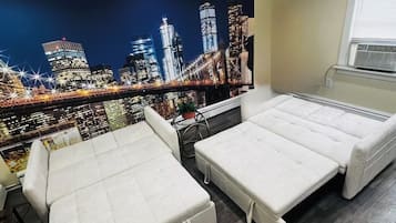 A room with two white beds, a window with blinds, a radiator, and a cityscape mural on the wall.